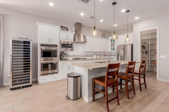kitchen with wine cooler, an island with sink, wall chimney range hood, a sink, and appliances with stainless steel finishes
