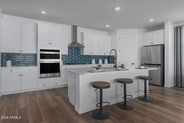 kitchen with an island with sink, stainless steel appliances, dark wood-type flooring, and wall chimney range hood