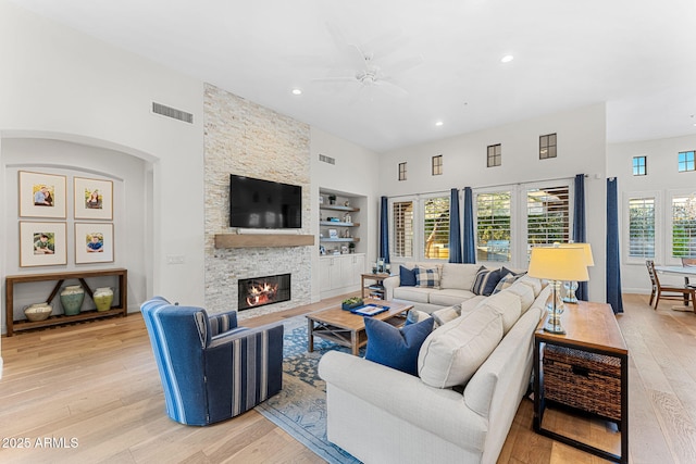 living room with a towering ceiling, built in features, a fireplace, ceiling fan, and light hardwood / wood-style floors