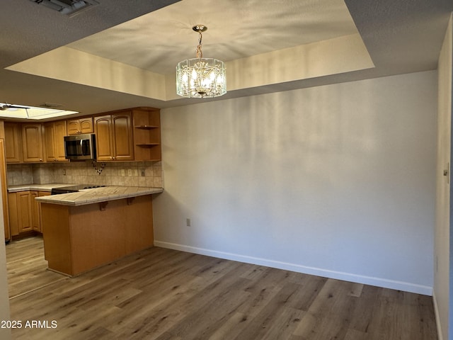 kitchen with light wood finished floors, a peninsula, a tray ceiling, stainless steel microwave, and tasteful backsplash