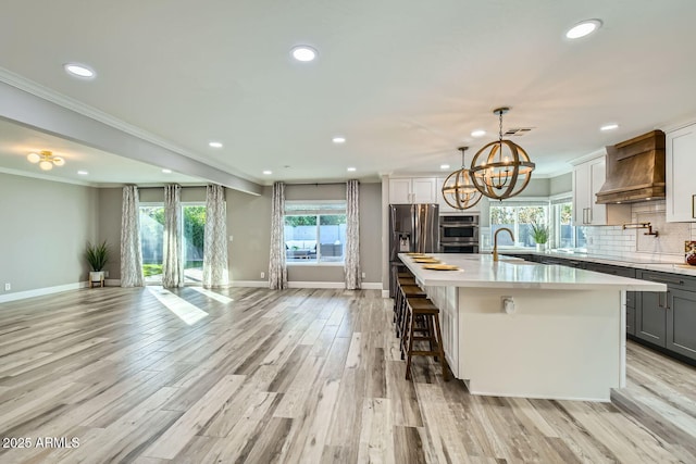 kitchen with ornamental molding, custom exhaust hood, an island with sink, and light wood-type flooring