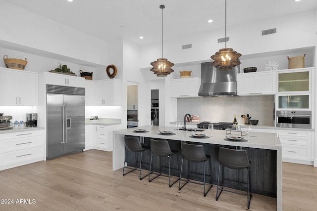 kitchen with appliances with stainless steel finishes, wall chimney exhaust hood, a breakfast bar, a center island with sink, and light hardwood / wood-style floors