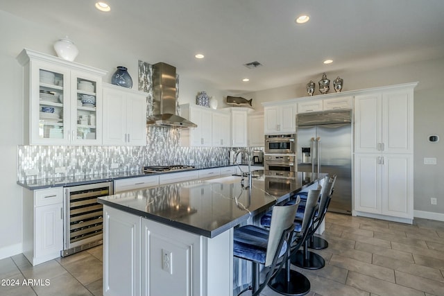 kitchen with appliances with stainless steel finishes, a kitchen island with sink, wall chimney range hood, white cabinets, and wine cooler