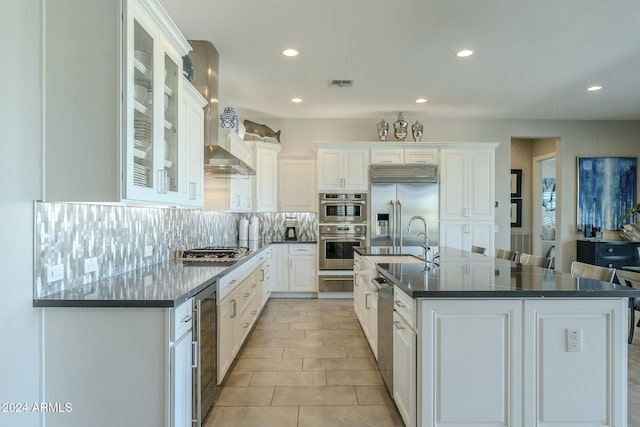 kitchen with wall chimney range hood, an island with sink, decorative backsplash, white cabinets, and appliances with stainless steel finishes