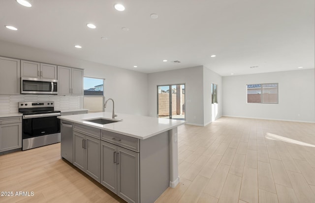 kitchen with gray cabinetry, sink, stainless steel appliances, backsplash, and an island with sink