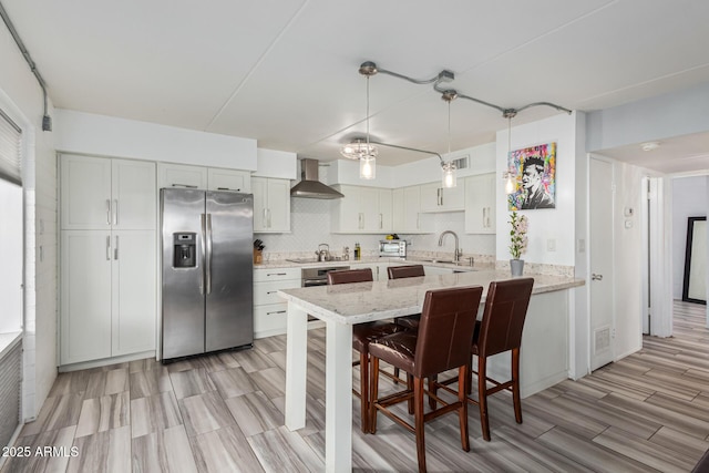 kitchen with white cabinetry, stainless steel fridge with ice dispenser, kitchen peninsula, light stone countertops, and wall chimney range hood