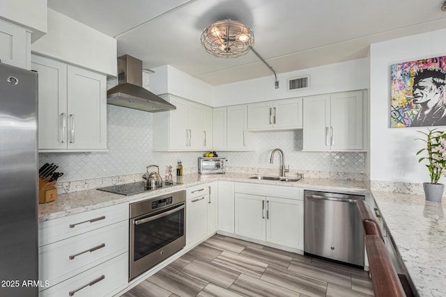 kitchen with sink, wall chimney range hood, stainless steel appliances, light stone counters, and white cabinets