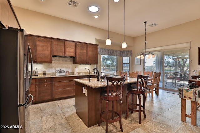 kitchen featuring sink, stainless steel refrigerator, light stone countertops, a center island with sink, and decorative backsplash