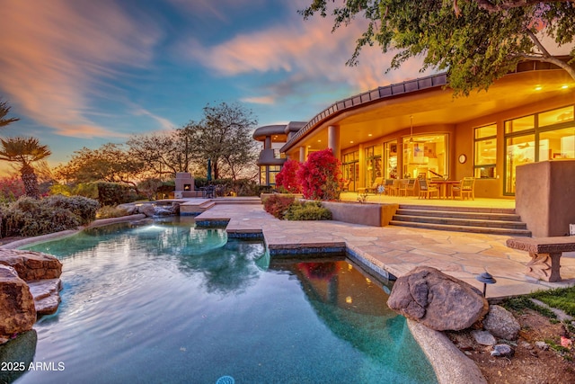 pool at dusk with pool water feature, a patio area, and an outdoor stone fireplace