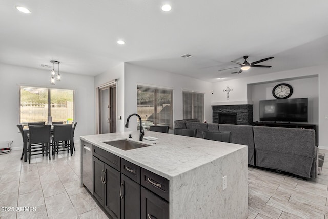 kitchen with dishwasher, a kitchen island with sink, sink, ceiling fan, and a fireplace