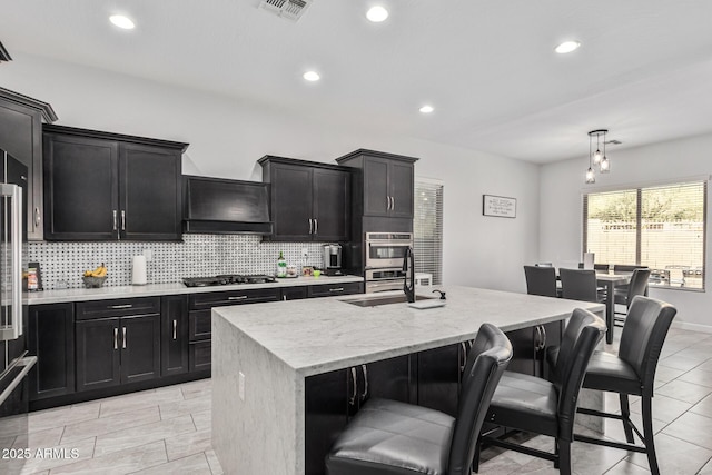 kitchen featuring backsplash, custom range hood, sink, pendant lighting, and a center island with sink