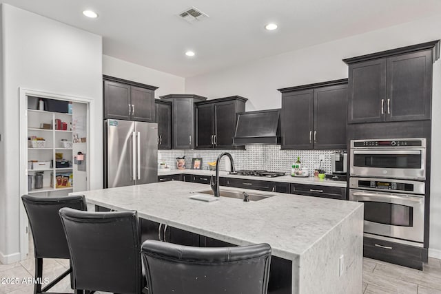 kitchen with premium range hood, a kitchen island with sink, sink, light stone countertops, and stainless steel appliances