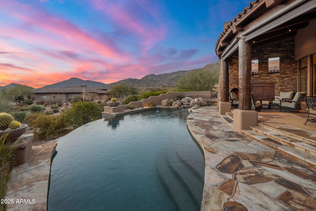 pool at dusk featuring a mountain view, a patio area, and an outdoor stone fireplace