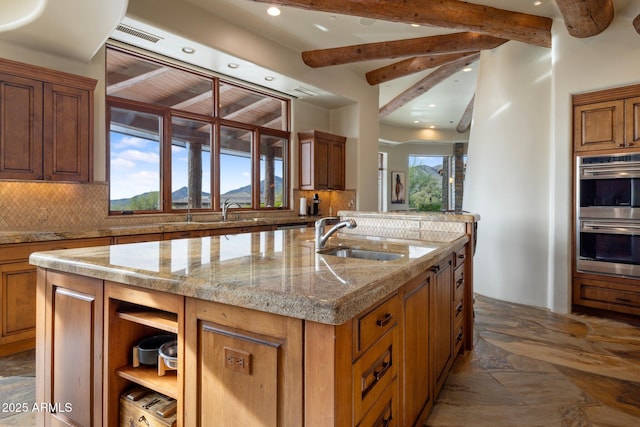 kitchen with sink, tasteful backsplash, light stone counters, an island with sink, and stainless steel double oven