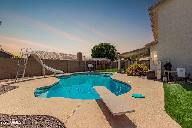 pool at dusk with a fenced backyard, a fenced in pool, a patio area, a diving board, and a water slide