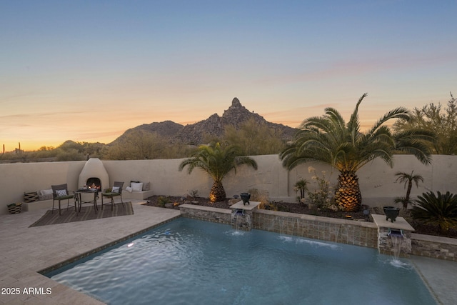 pool at dusk featuring exterior fireplace, pool water feature, a mountain view, and a patio