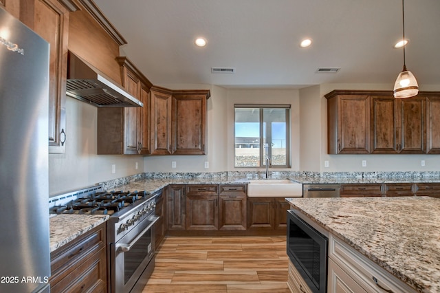 kitchen featuring visible vents, a sink, light stone counters, stainless steel appliances, and wall chimney exhaust hood