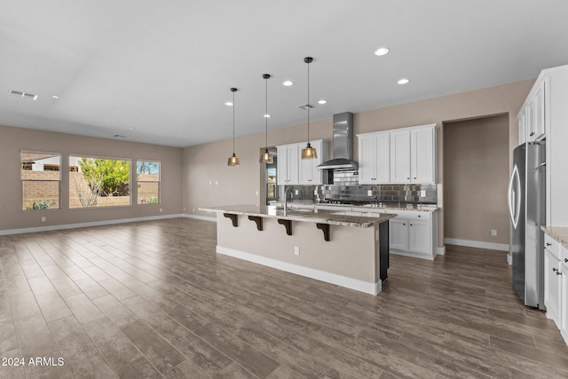 kitchen with white cabinetry, a kitchen island with sink, stainless steel refrigerator, and wall chimney range hood