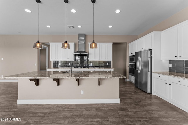 kitchen featuring hanging light fixtures, wall chimney range hood, an island with sink, white cabinets, and appliances with stainless steel finishes