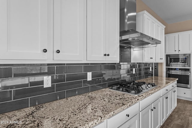 kitchen featuring wall chimney exhaust hood, tasteful backsplash, light stone counters, white cabinetry, and stainless steel appliances