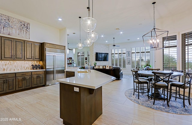 kitchen featuring an island with sink, a sink, stainless steel built in fridge, light countertops, and tasteful backsplash