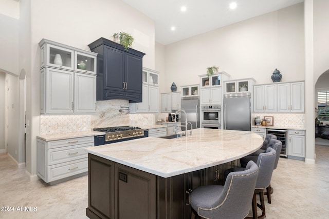 kitchen with stainless steel appliances, wine cooler, a towering ceiling, a kitchen bar, and a center island with sink