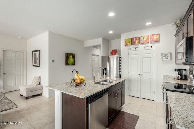 kitchen with an island with sink, sink, stainless steel appliances, light stone countertops, and dark brown cabinets