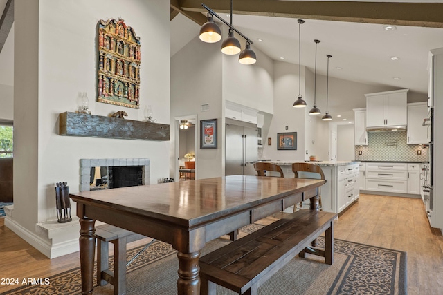 dining room with a tiled fireplace, light wood-type flooring, beam ceiling, and high vaulted ceiling