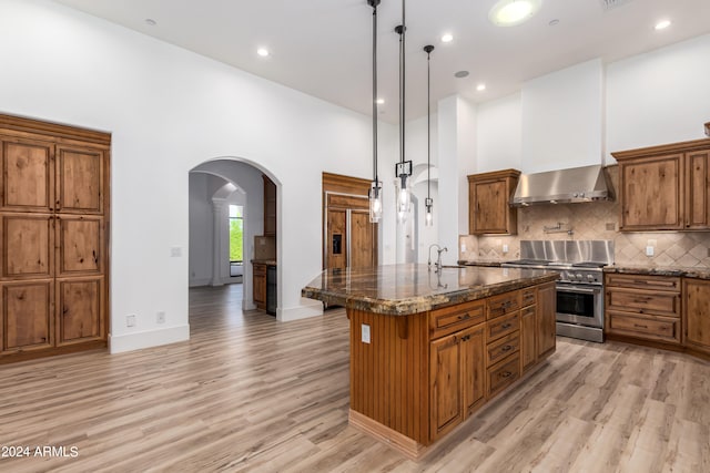 kitchen with wall chimney range hood, hanging light fixtures, an island with sink, a high ceiling, and stainless steel range