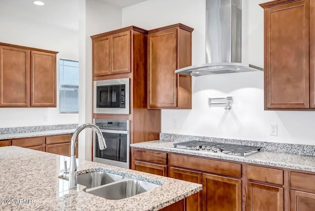 kitchen featuring a sink, brown cabinets, stainless steel appliances, wall chimney exhaust hood, and light stone counters