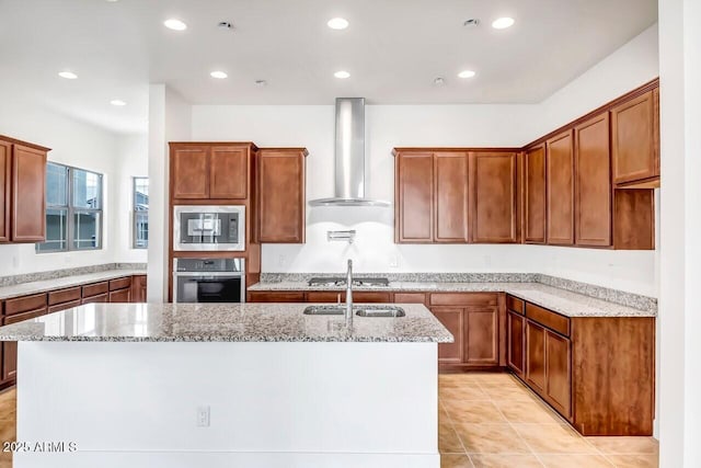 kitchen featuring light stone countertops, a kitchen island with sink, appliances with stainless steel finishes, a sink, and wall chimney exhaust hood