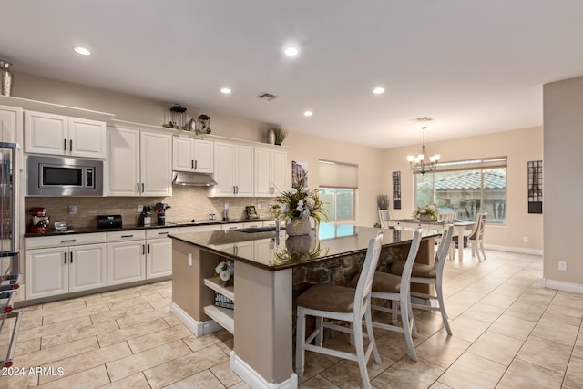 kitchen featuring white cabinetry, stainless steel microwave, a kitchen island with sink, an inviting chandelier, and a breakfast bar area