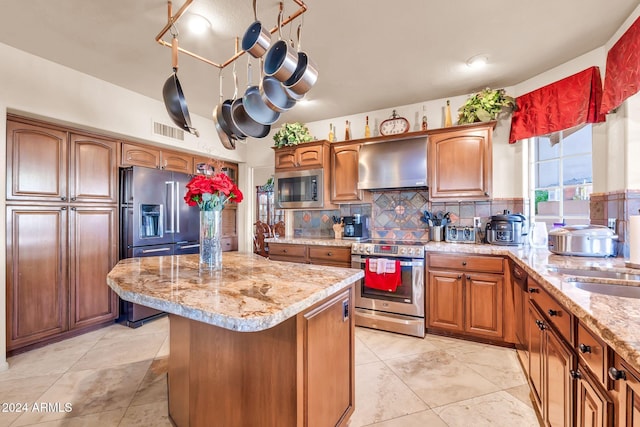 kitchen featuring wall chimney exhaust hood, appliances with stainless steel finishes, a kitchen island, light stone countertops, and backsplash