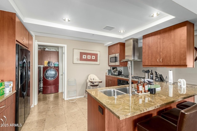 kitchen with black refrigerator with ice dispenser, wall chimney exhaust hood, a raised ceiling, brown cabinetry, and baseboards