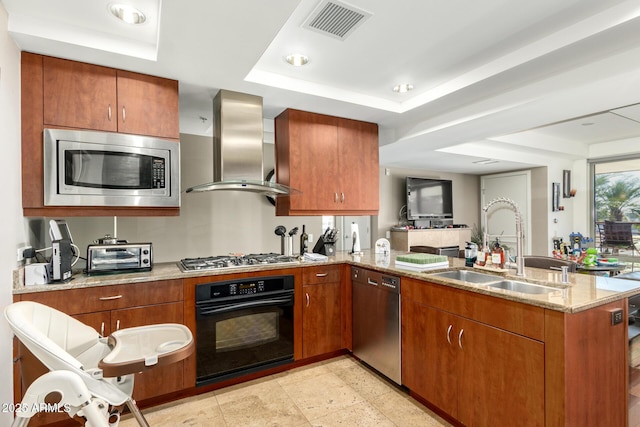 kitchen featuring stainless steel appliances, wall chimney range hood, a raised ceiling, a sink, and a peninsula