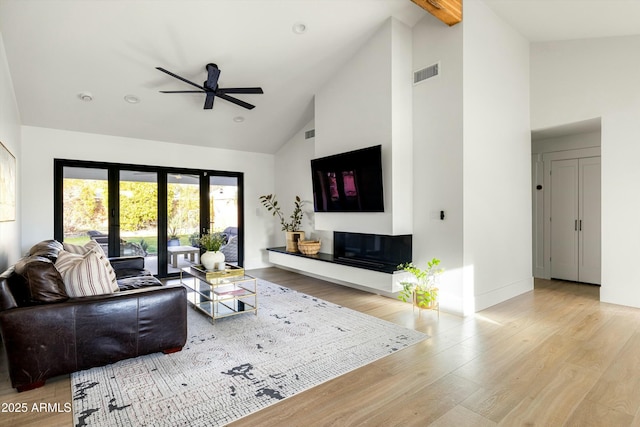 living area featuring ceiling fan, high vaulted ceiling, wood finished floors, visible vents, and a glass covered fireplace