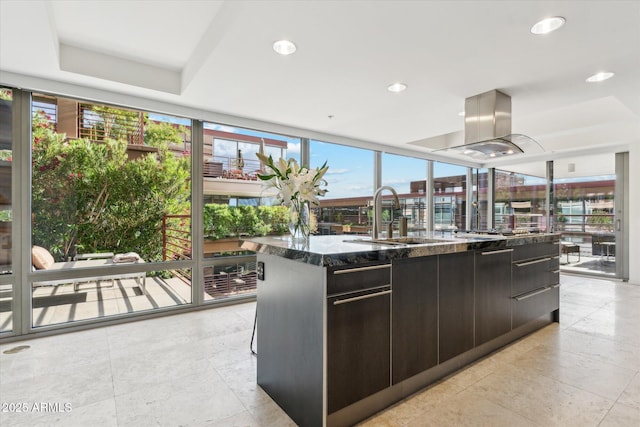 kitchen with a sink, dark stone countertops, island range hood, floor to ceiling windows, and a center island with sink