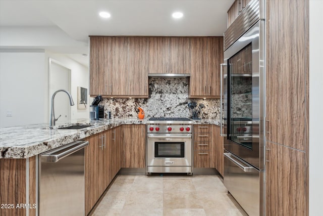 kitchen featuring light stone countertops, brown cabinets, a sink, a peninsula, and premium appliances