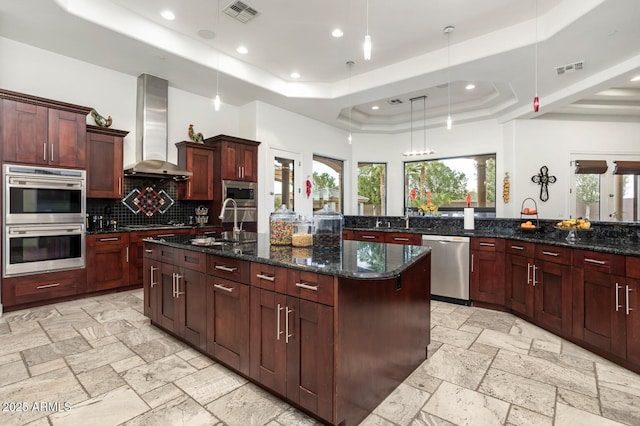 kitchen featuring a large island with sink, pendant lighting, a raised ceiling, stainless steel appliances, and wall chimney exhaust hood