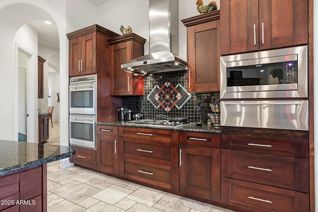 kitchen with appliances with stainless steel finishes, decorative backsplash, wall chimney exhaust hood, and dark stone counters