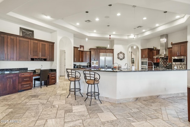 kitchen featuring a high ceiling, hanging light fixtures, built in appliances, a large island with sink, and wall chimney range hood