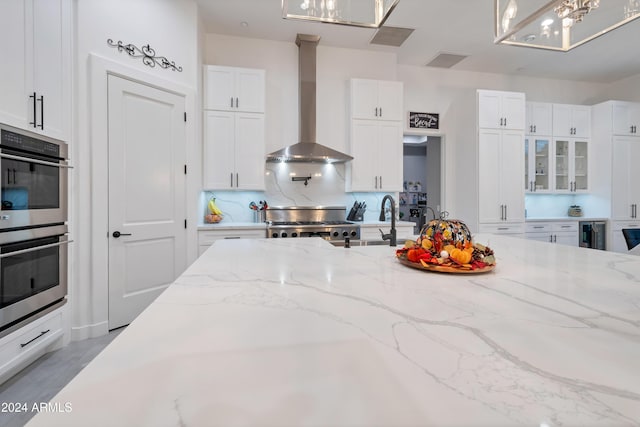 kitchen featuring stove, wall chimney range hood, double oven, light stone counters, and white cabinetry