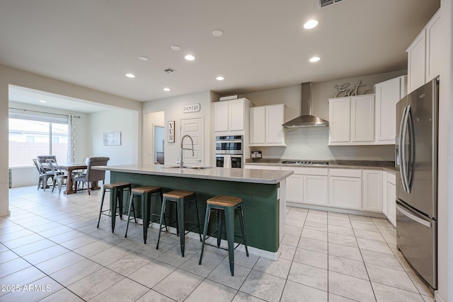 kitchen with appliances with stainless steel finishes, wall chimney range hood, a kitchen island with sink, and white cabinets