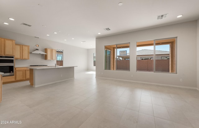 kitchen with an island with sink, stovetop, light brown cabinetry, double oven, and sink