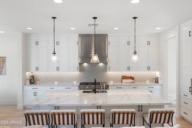 kitchen featuring white cabinets, light stone counters, wall chimney exhaust hood, and an island with sink