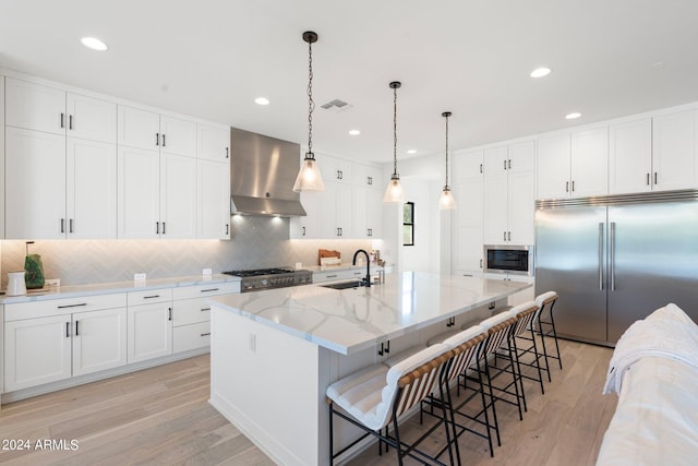 kitchen with light stone counters, wall chimney exhaust hood, built in appliances, white cabinets, and an island with sink