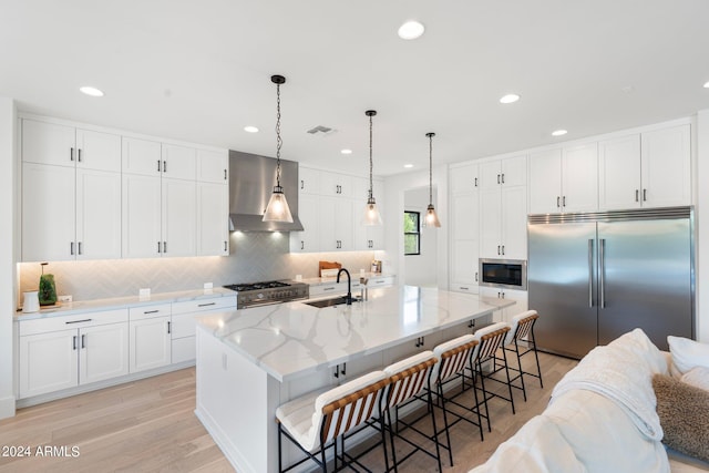 kitchen with a kitchen island with sink, wall chimney range hood, built in appliances, white cabinetry, and a breakfast bar area