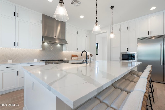 kitchen with built in appliances, wall chimney range hood, sink, and a large island with sink