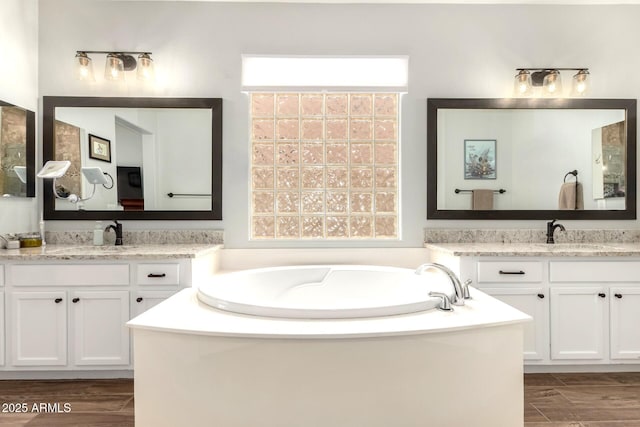 full bath with a sink, two vanities, a bath, and wood finish floors