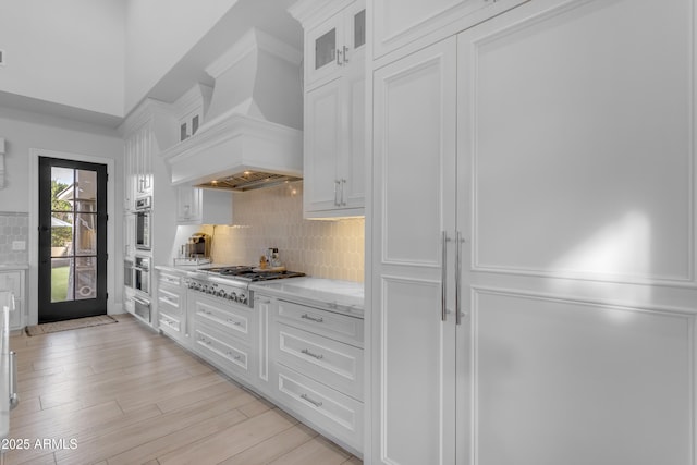 kitchen featuring light wood finished floors, white cabinets, decorative backsplash, appliances with stainless steel finishes, and custom exhaust hood